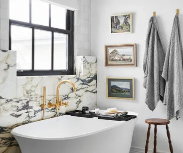 Luxurious marble bathroom with a freestanding bathtub, gold fixtures, paintings, and striped towels on wall hooks.