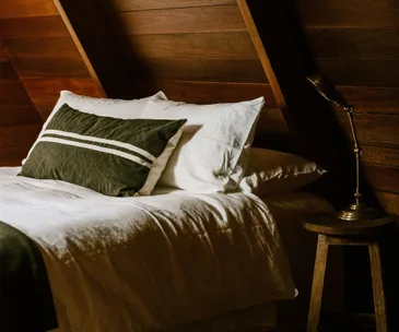 Cozy attic bedroom with a green pillow, white bedding, wooden walls, and a vintage lamp on a rustic stool.