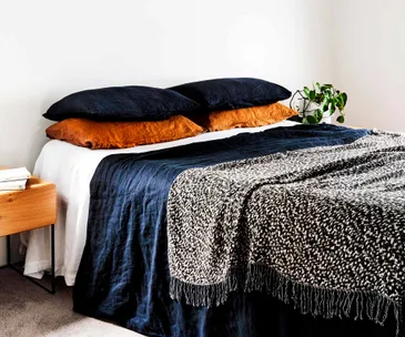 Neatly made bed with black and orange pillows, a bedside table, and a plant next to it.