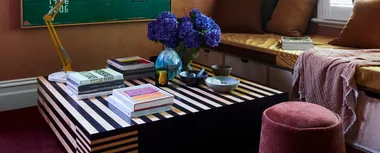 A jewel-toned sitting room with built-in bench seat, a large stiped coffee tables and plum-coloured carpet.