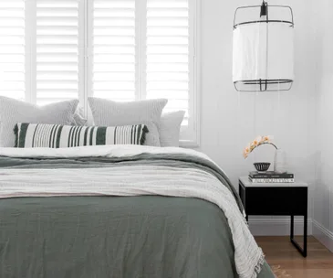 Modern bedroom with green bedding, striped pillows, white shutters, black nightstand, books, vase with flowers, and hanging lamp.