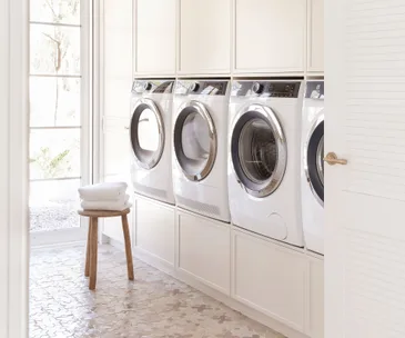 A modern laundry room with four front-load washers, white cabinets, and folded towels on a wooden stool.