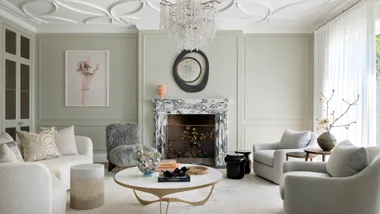 A living room with chandelier, two grey chairs, cream sofa, marble fireplace, artwork and contemporary mirror