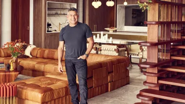 Jamie Durie in his Avalon house for sale wearing a black t shirt and jeans standing in his living room smiling.