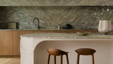 Modern kitchen with green marble backsplash, wooden cabinets, two wooden stools, and a large vase on the counter.