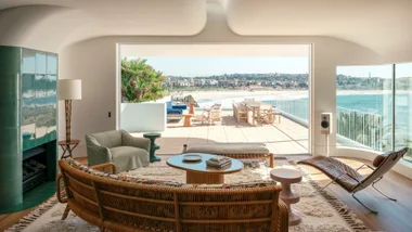 Living room with wicker furniture overlooking a seaside balcony with dining set and scenic coastal view.