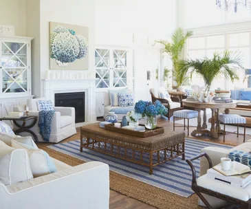 Bright living room with white furniture, blue accents, wicker coffee table, large windows, and a painting of hydrangeas above the fireplace.