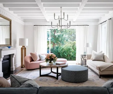 Elegant living room with white beamed ceiling, large windows, pink sofa, round ottoman, cushioned chairs, and a chandelier.