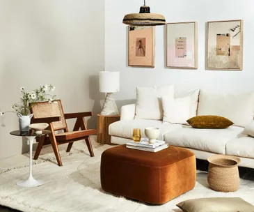 Neutral-toned living room with a white sofa, wooden chair, brown ottoman, side table, plants, table lamp, and wall art.