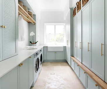 Spacious laundry room with light blue cabinets, built-in washer and dryer, wooden bench, wicker baskets, and a large window.