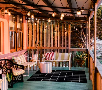 Cozy porch with string lights, patterned cushions, black and white rug, potted plants, and wooden slatted fence.