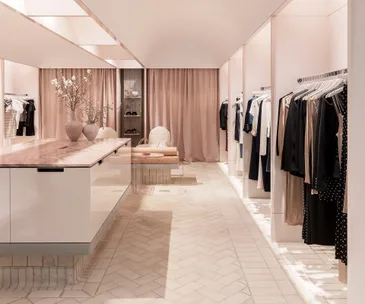 Minimalistic boutique with pink curtains, cream tiled floor, clothes on racks, a stone table with vases, and pastel seating area.