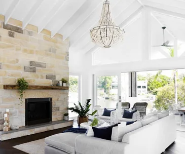 Bright living room with stone fireplace, gray sofa, chandelier, and large windows overlooking a patio and greenery.