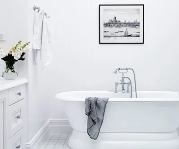 Clean white bathroom with a freestanding tub, gray towel, white cabinets, flowers on the vanity, and a framed picture on the wall.