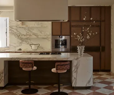 Luxurious kitchen with marble countertops, two pink bar stools, wood cabinets, and a vase with branches on the island.