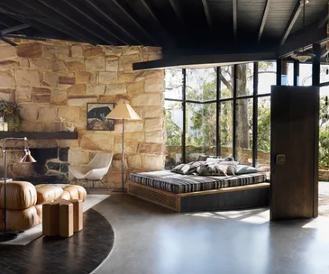 Stone-walled living room with large windows, brown leather sofa, fireplace, striped bed, and natural outdoor view.