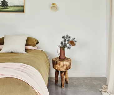 Simple, modern bedroom with a brown bedspread, white pillow, and wooden nightstand holding a vase with greenery.
