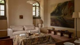 A living room with a grey linen couch, artwork of natural landscape, pattern fabric coffee table, terracotta brickwork flooring and frosted arched windows with grey frames.