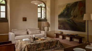 A living room with a grey linen couch, artwork of natural landscape, pattern fabric coffee table, terracotta brickwork flooring and frosted arched windows with grey frames.