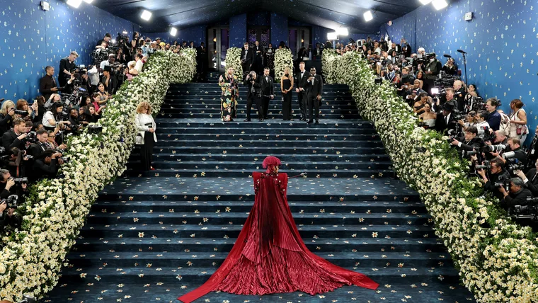 A blue staired carpet with flowers ad woman in a red dress and hat for met gala theme article