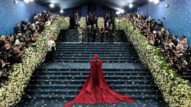 A blue staired carpet with flowers ad woman in a red dress and hat for met gala theme article