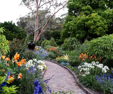 Brick path winds through garden with flower beds full of orange, blue and white flowers with trees behind