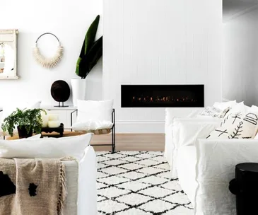 A modern living room with a wall-mounted gas fireplace, white furniture, plants, and a geometric rug.