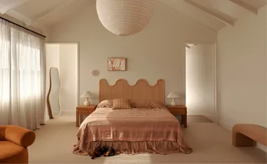 Cozy bedroom with wicker headboard, soft bedding, two lamps, a mirror, and a dog sleeping on the floor.