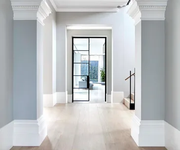 Open hallway with light timber flooring, grey walls, decorative columns, and glass doors leading to a bright living area and garden.