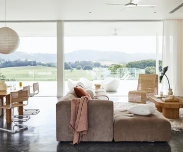 Modern living room with large windows, a brown couch, wicker furniture, and views of a green landscape and hills in the distance.