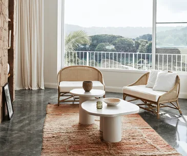 Bright living room with rattan chairs, white cushions, round tables, and a large window overlooking greenery.